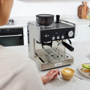 Kvinna njuter cappuccino bredvid espressomaskin och avokadotoast i modernt kök.