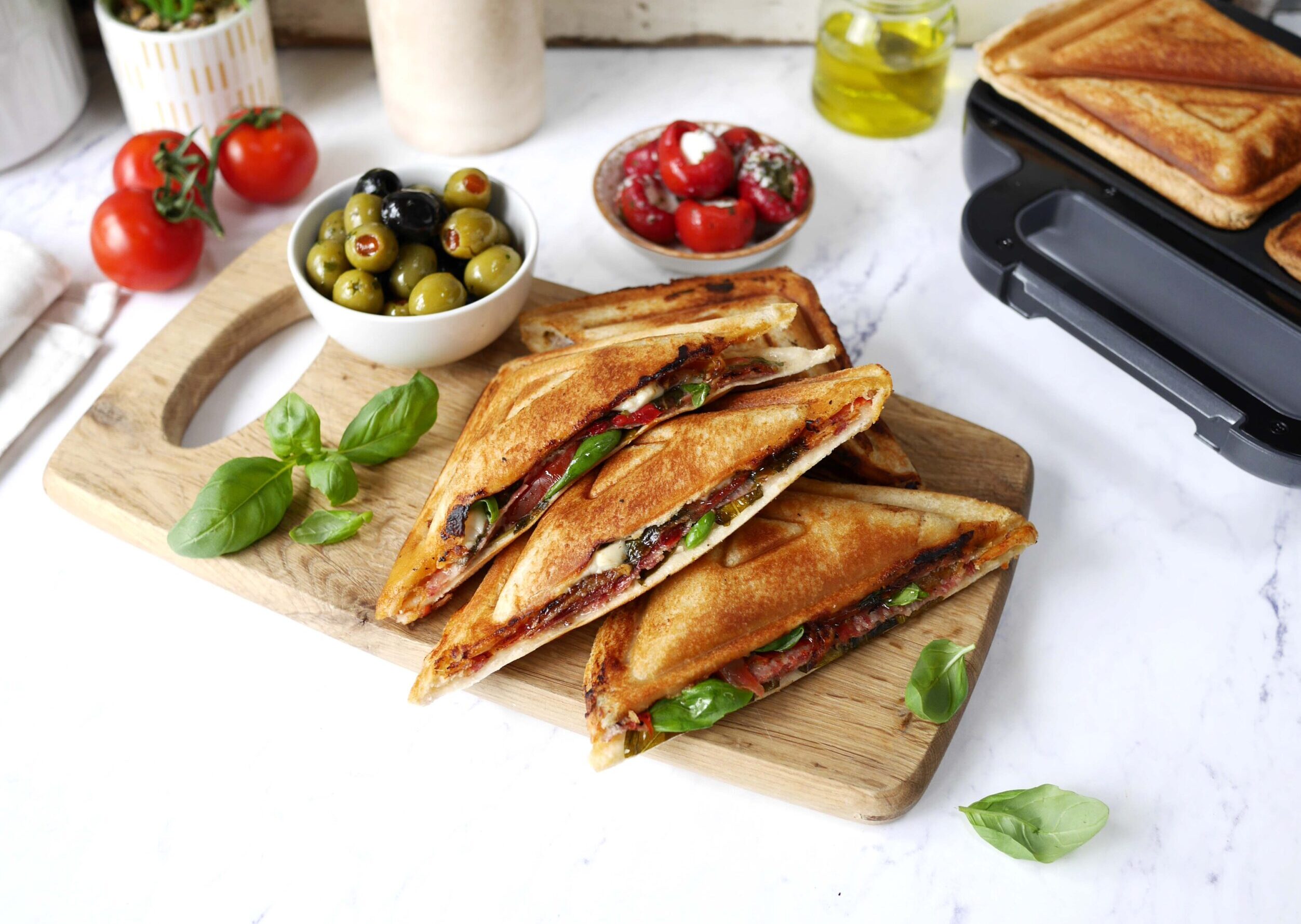 Grillade smörgåsar med basilika, oliver och tomater på en träbricka i kök.